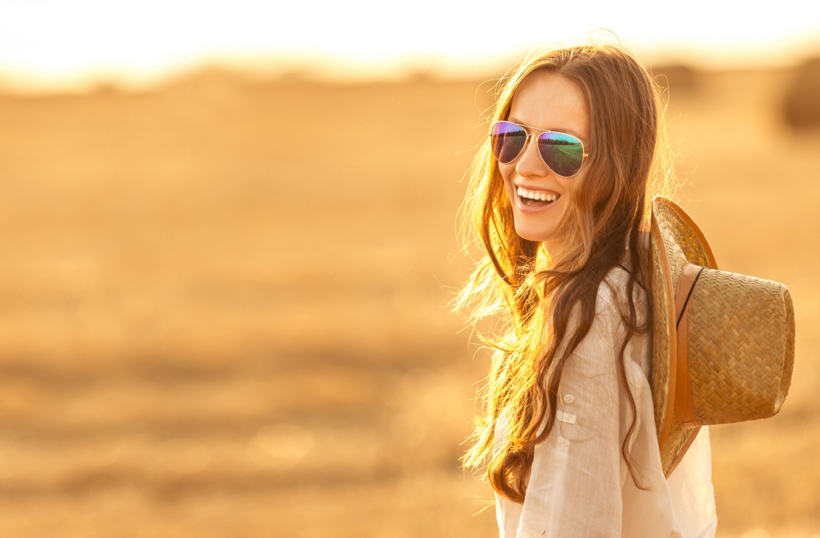 A person wearing polarized sunglasses laughs as the sun sets behind them. A straw hat sits on their back between their shoulders.